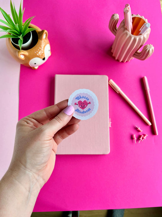 Hand holding a sticker with a pink heart design over a pink notebook on a pink background.