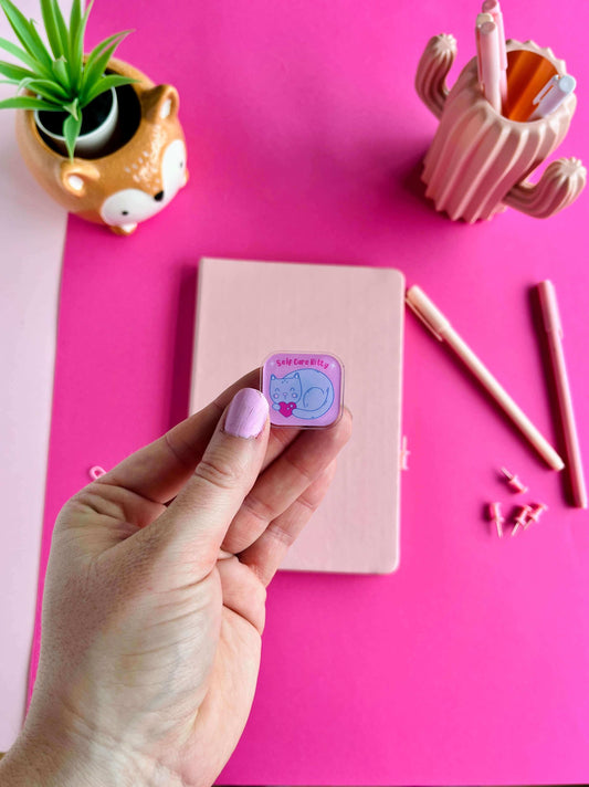 Hand holding a small pink and purple enamel pin with a cat design on a pink surface with stationery items.